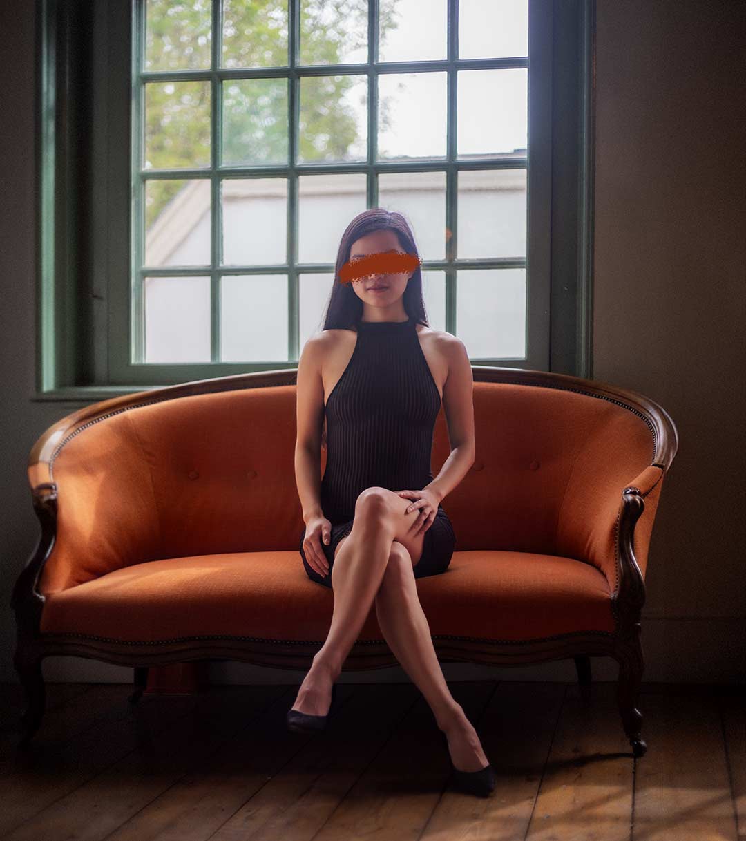 A woman in a sleeveless black dress sits elegantly on an orange vintage sofa placed in front of a large window with green views. Her legs are crossed, and she wears black high heels. The room has a rustic charm with dark green walls and a wooden floor, illuminated by natural light coming through the window.